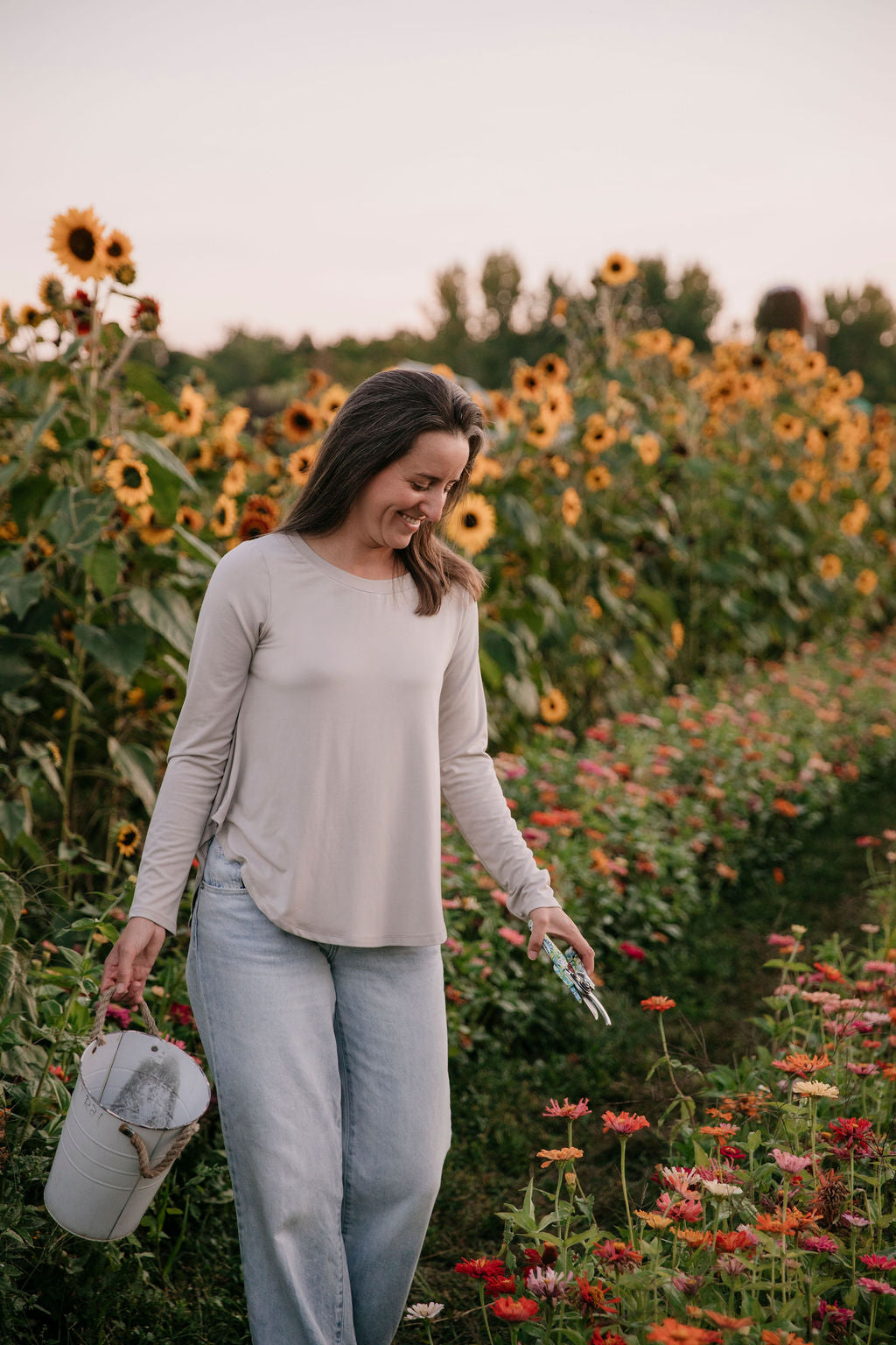 Woman wearing The Breezy Long-Sleeve Shirt in Taupe, standing in a field of sunflowers and other flowers, holding a white bucket and scissors.