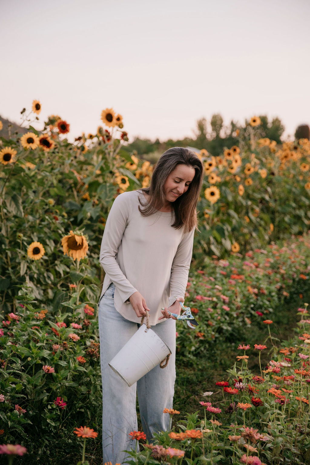 Woman wearing The Breezy Long-Sleeve Shirt in Taupe, standing in a field of sunflowers and other flowers, holding a white bucket.