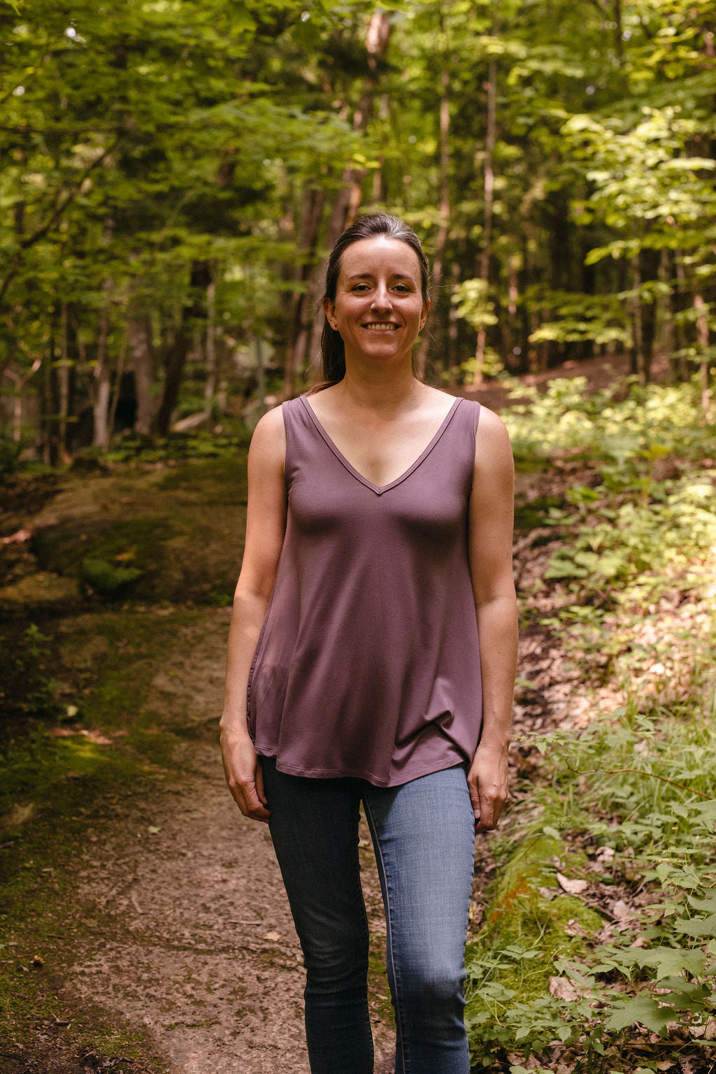 Woman standing on a forest path wearing a lavender sleeveless top and blue jeans.