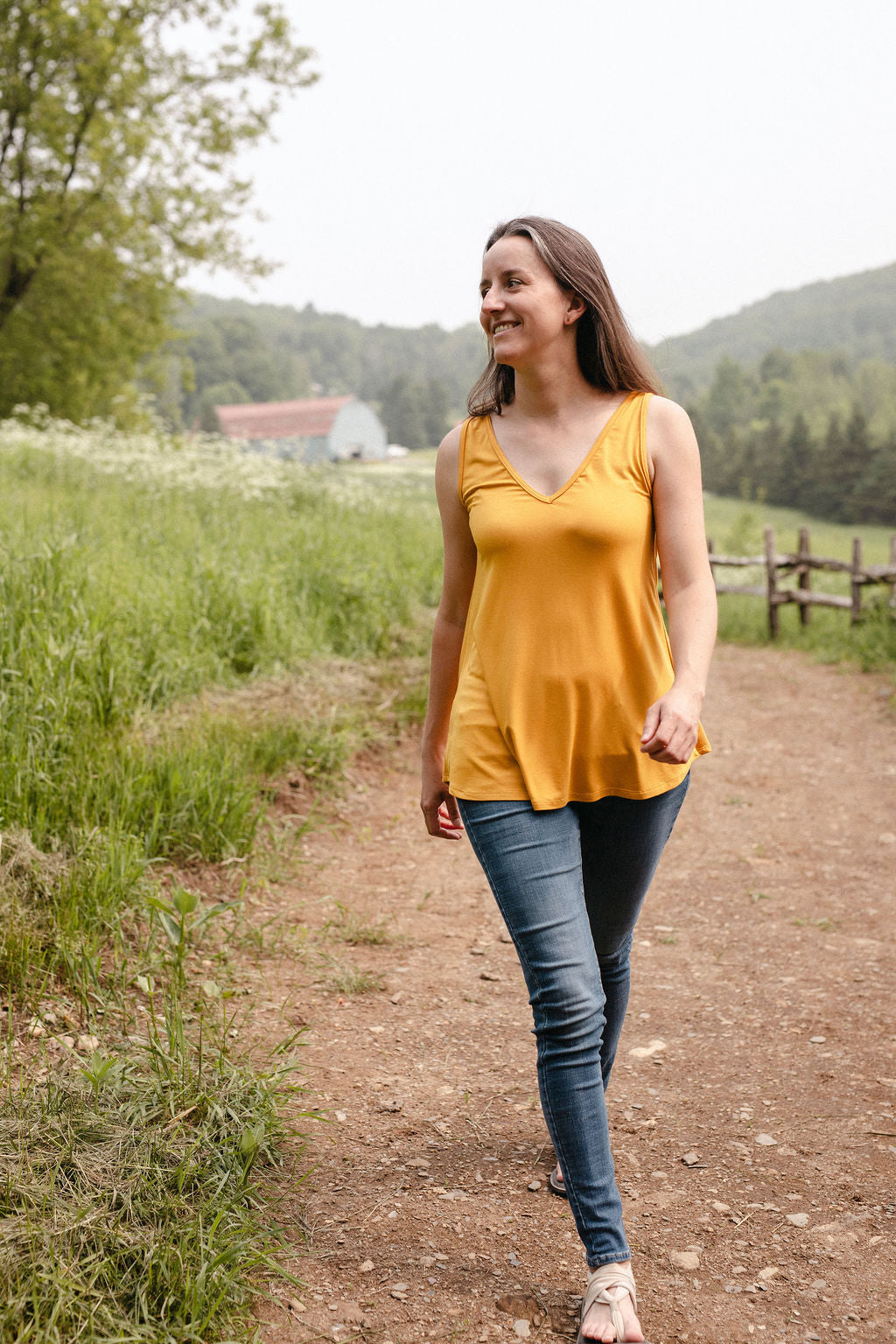 Woman in a yellow sleeveless top and blue jeans walking on a dirt path in a scenic outdoor setting.