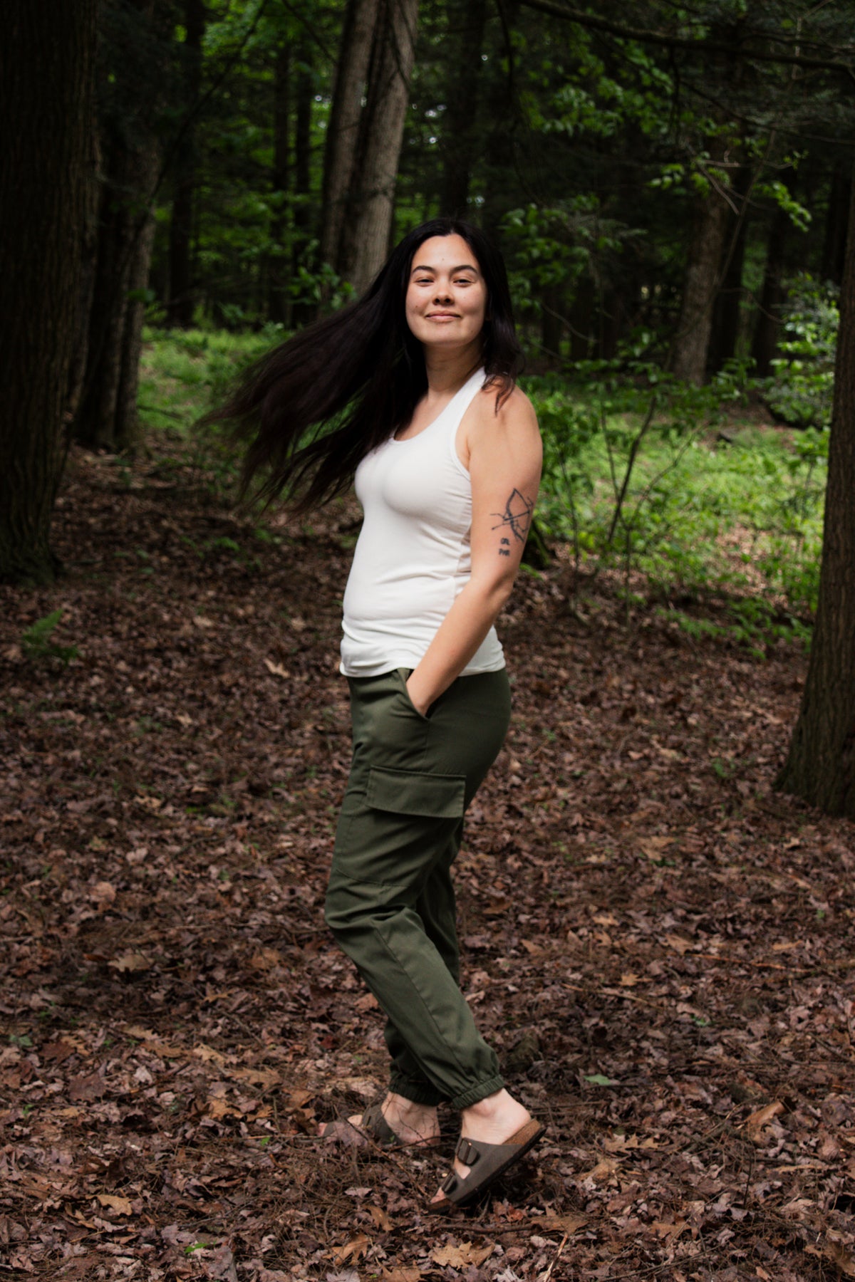 Woman standing in a forest wearing a white top and green pants.
