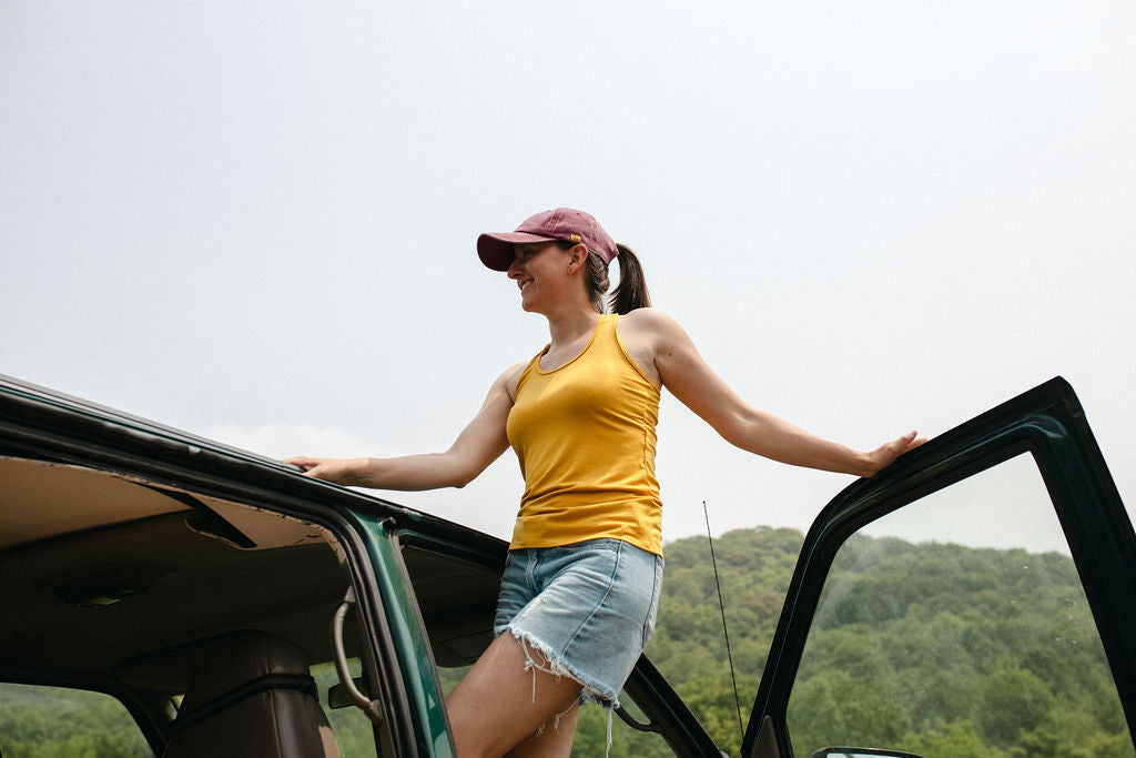 Woman in yellow tank top and denim shorts standing by an open car door with a scenic background.