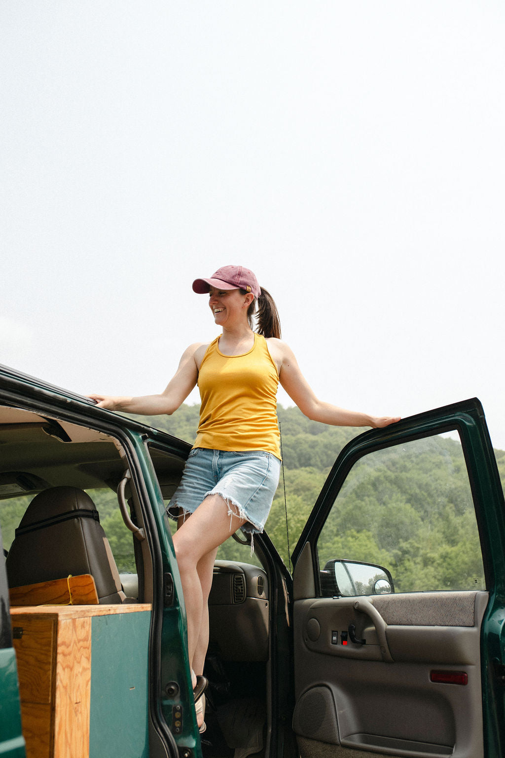 Woman in yellow tank top and pink cap getting out of a green camper van with a scenic background