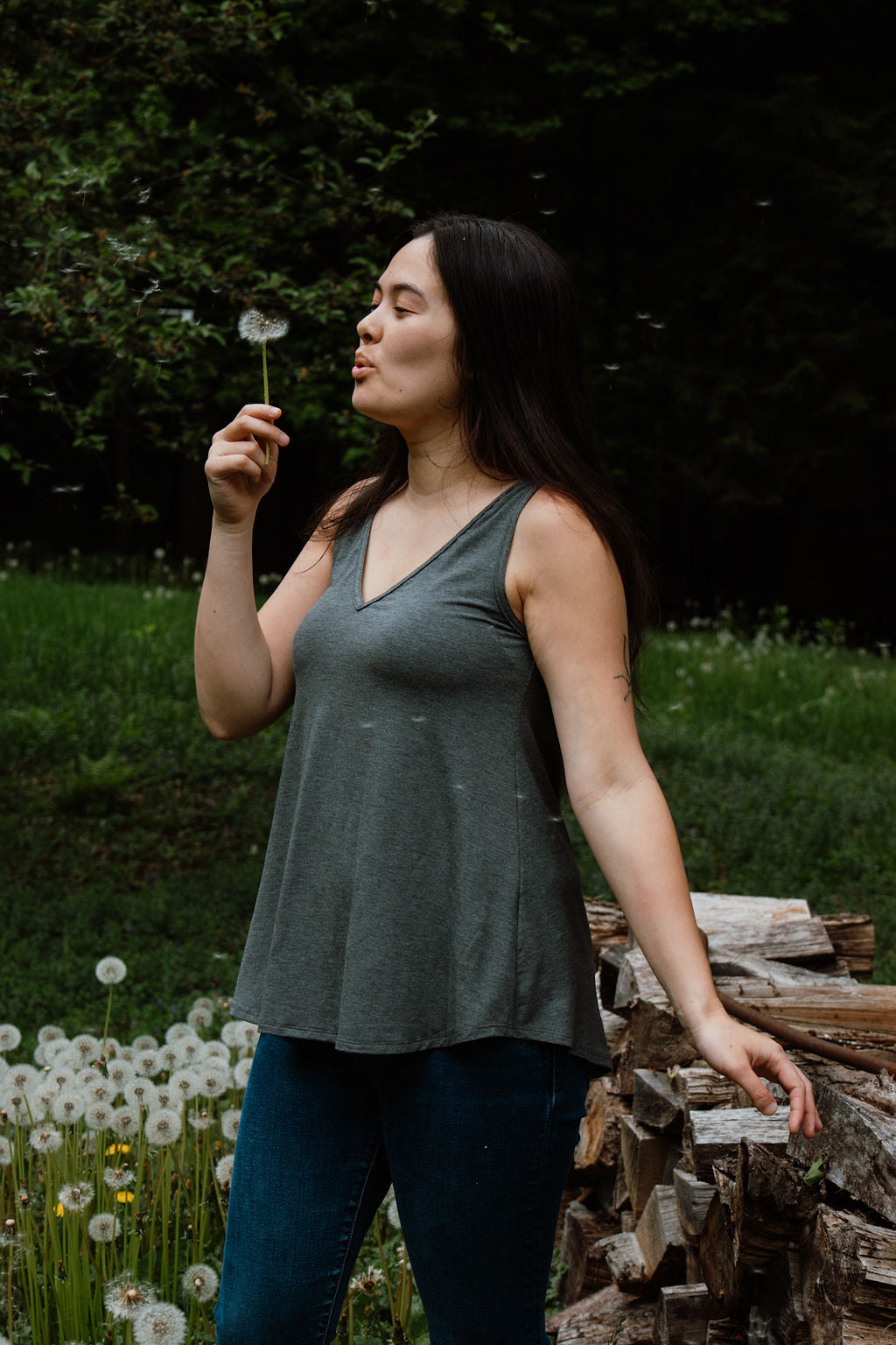 Woman blowing on a dandelion in a natural setting with trees and grass.