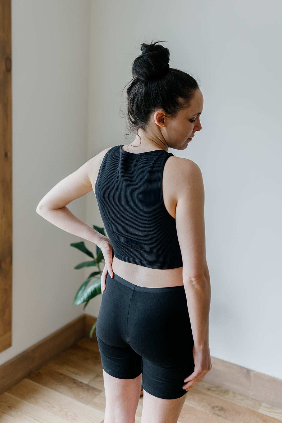 A woman wearing a black top and long bamboo boxer shorts, standing in a room with plants in the background