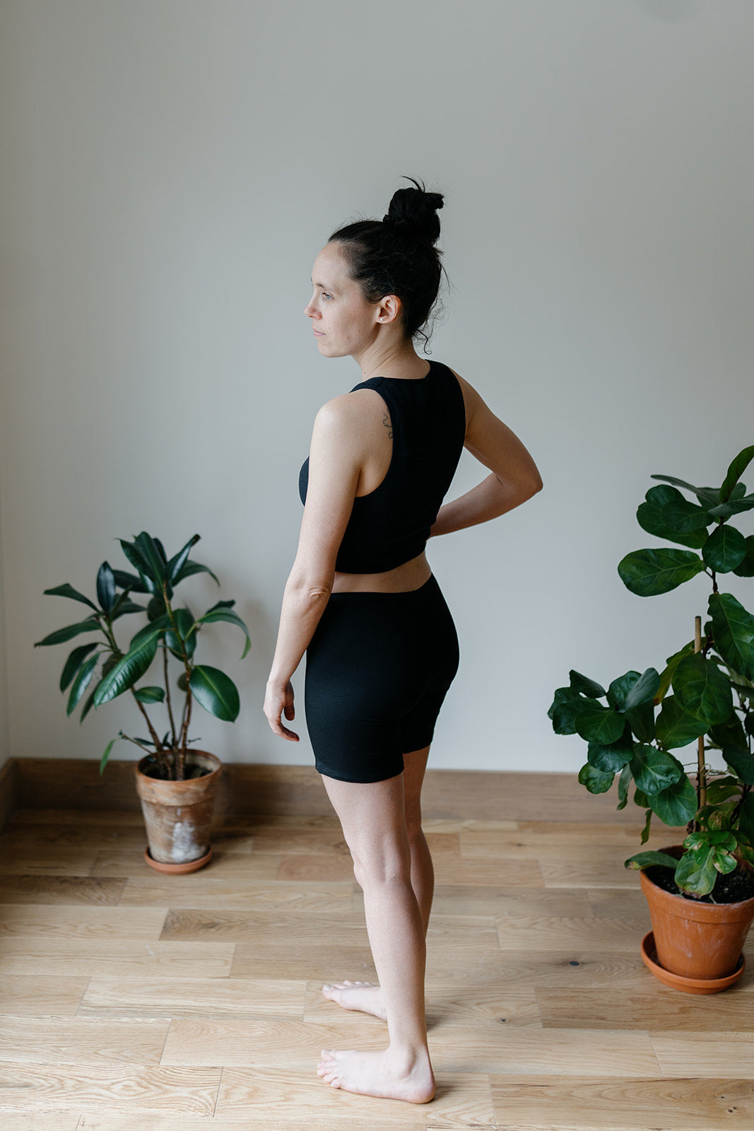 A woman wearing a black top and long bamboo boxer shorts, standing in a room with plants in the background