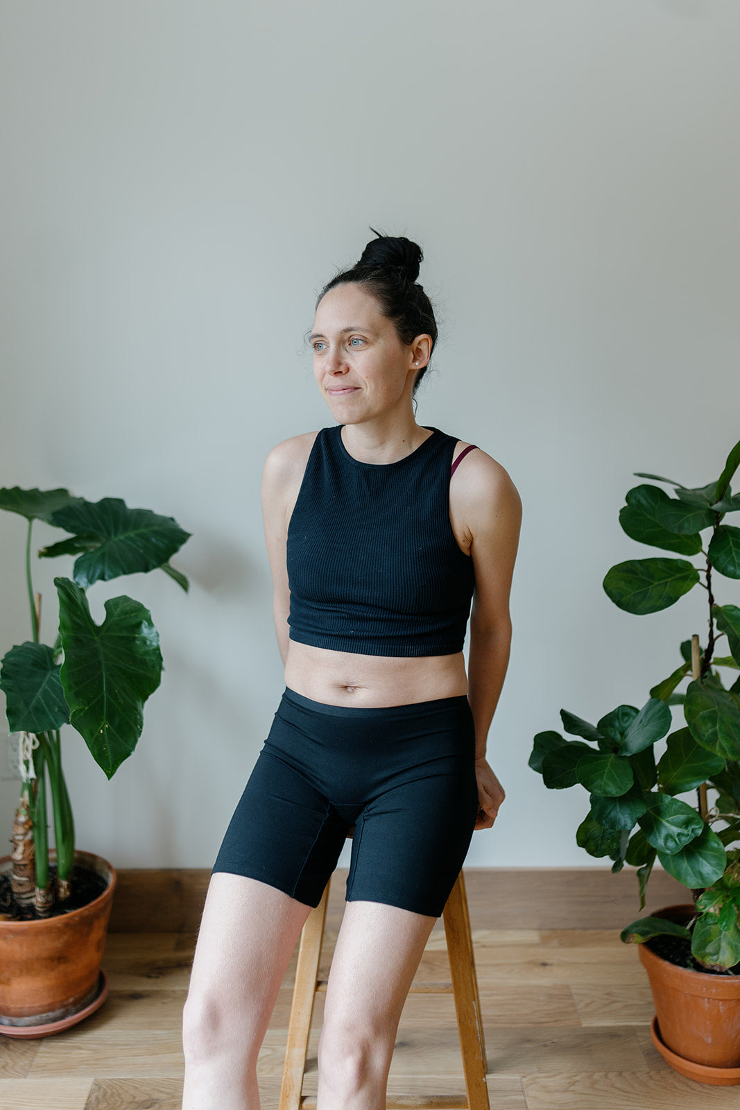 A woman wearing a black top and long bamboo boxer shorts, sitting on a stool with plants in the background