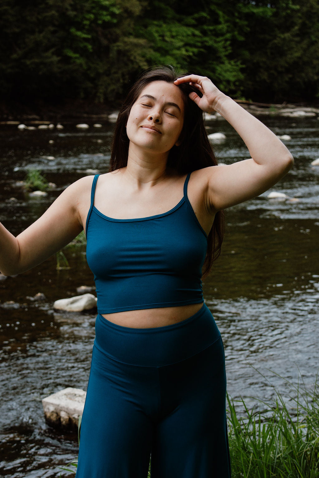 Woman wearing a zero-waste crop top in teal with teal Palazzo pants standing in a grassy field in front of a river.