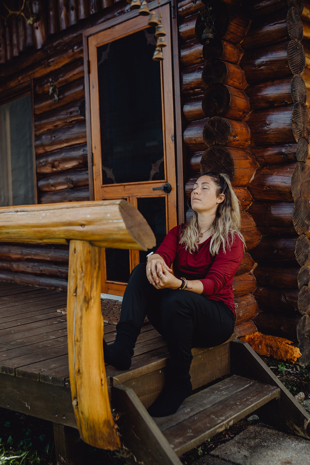 A person sitting on wooden steps leading to a cabin door, wearing black pants, a red top and black wool socks