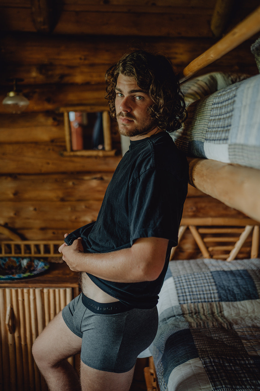 A man wearing grey trunks with a black shirt, standing in front of a bed.